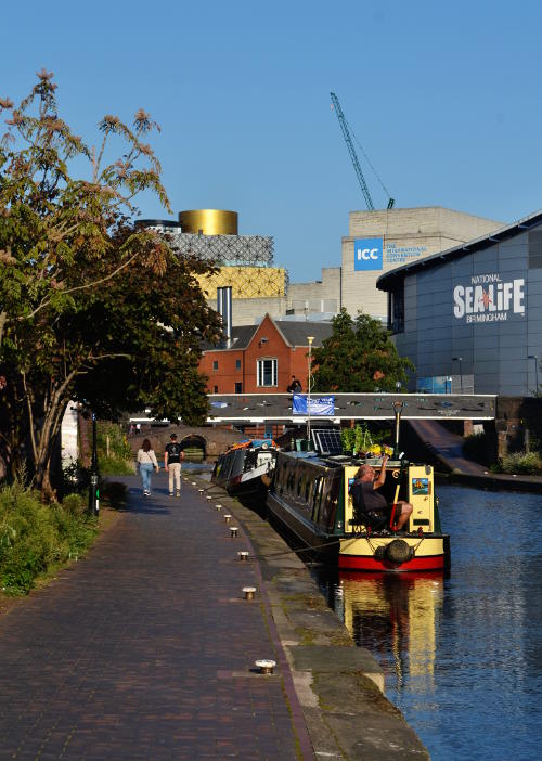 Walkers by a canal with the Library of Birmingham, the ICC and Sea Life Centre in the distance