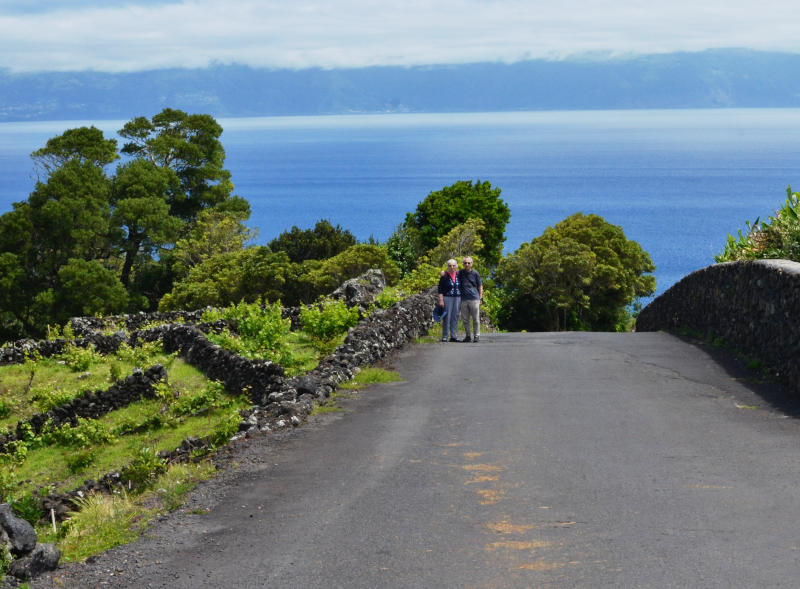 Phil and Miriam standing in a road with the sea behind and a small vineyard on the left