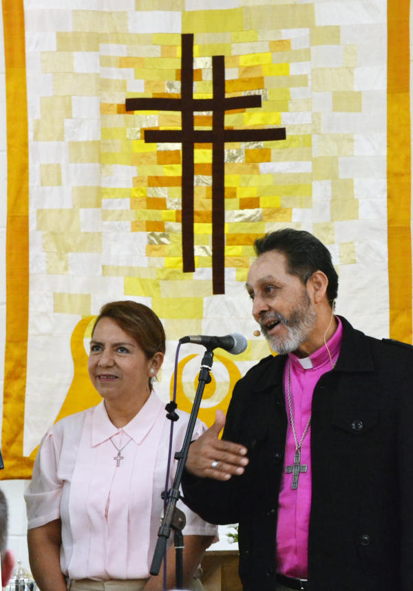 Walter speaks into a microphone with Adela at his side and a cross-themed wall hanging behind them