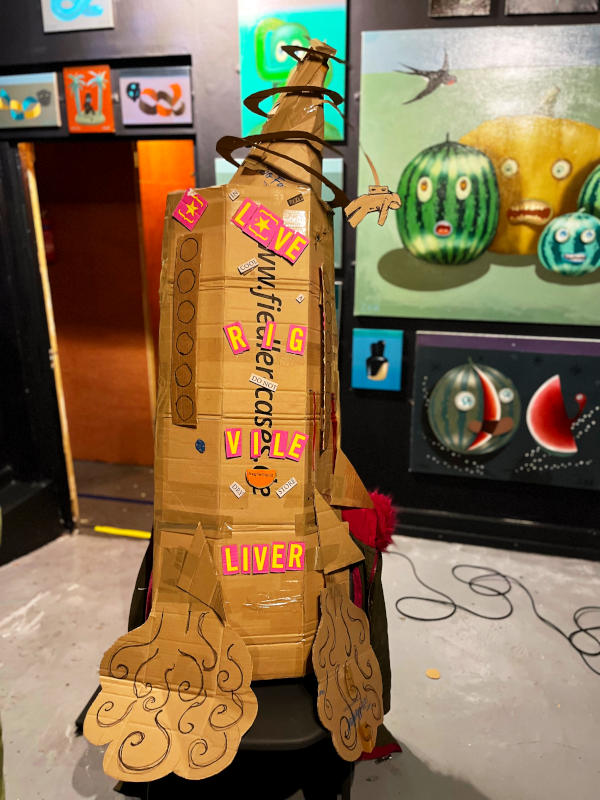 A cardboard rocket with an art exhibition behind