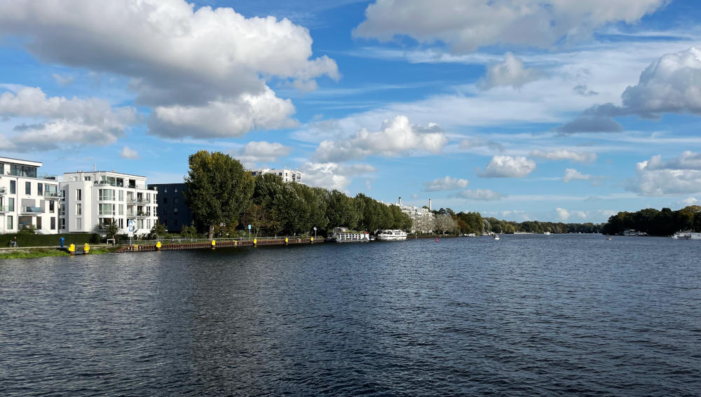 A broad stretch of water with trees and white buildings on the left bank