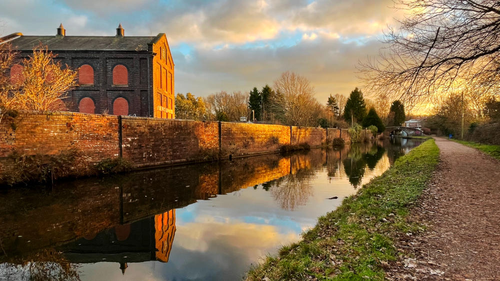 A historic building, wall and towpath along a canal, with a footbridge in the distance, in dramatic evening light
