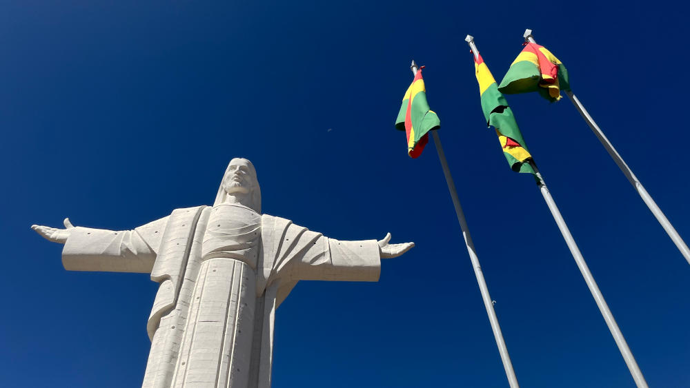 Looking up at a statue of Christ next to 3 Bolivian flags on flagpoles, with deep blue sky
