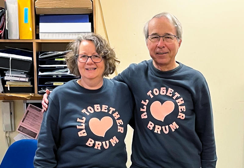 Amanda and Phil in an office wearing grey swetshirts each with a pink heart and the words "All Together Brum"
