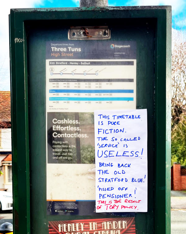 A bus stop with a handwritten notice complaining that the service is useless, signed by "Pissed Off Pensioner"