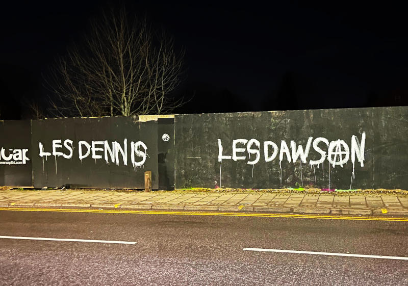 Graffiti in white on a black fence at night, under street lights: "Les Dennis" and "Les Dawson"