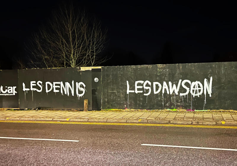 Graffiti in white on a black fence at night, under street lights: "Les Dennis" and "Les Dawson"