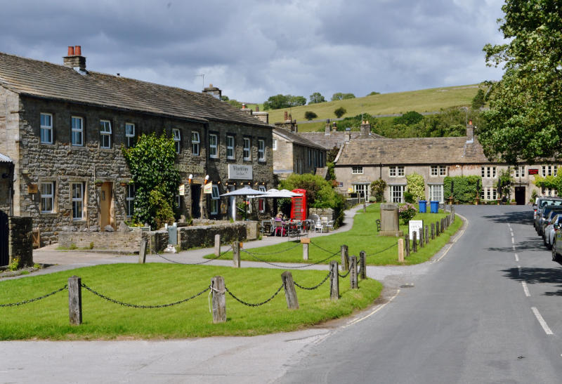 A row of cafés and shops built of stones with a wide grass verge and a road in front