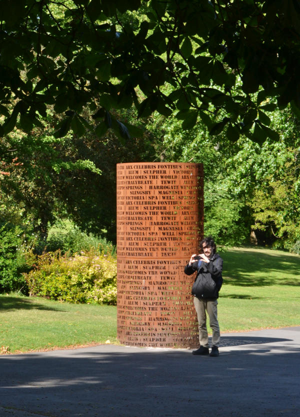 Sculpture consisting of a tall metal cylinder with stencilled letters hollowed out. Martin stands next to it, looking at his camera