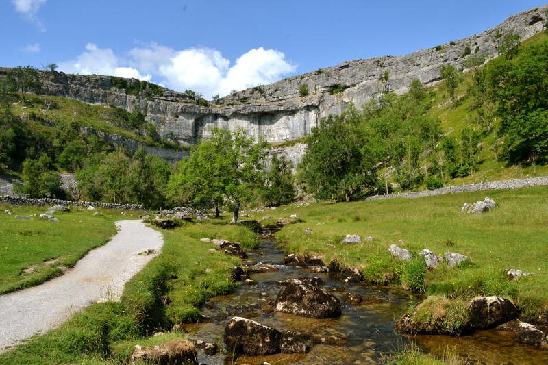 A high cliff face behind a stream flowing between a path and grassland