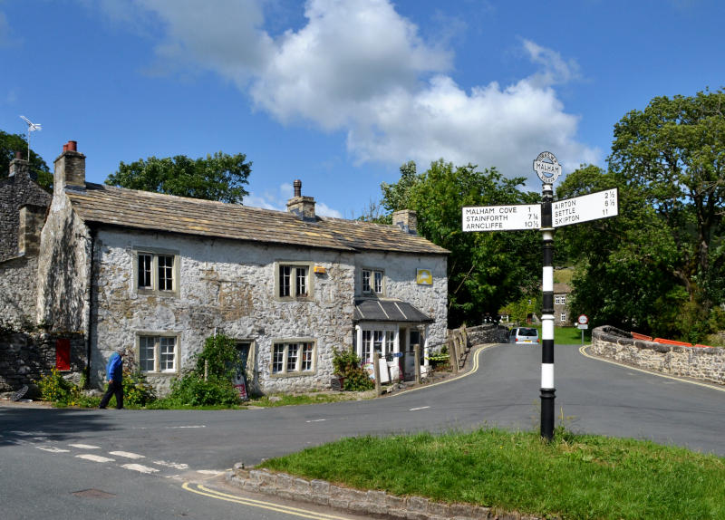 An old cottage-style building behind a road junction, with signs to Malham Cove, Settle and other places