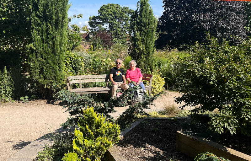 Phil and Miriam sitting on a wooden bench with trees and shrubs all around, in sunshine