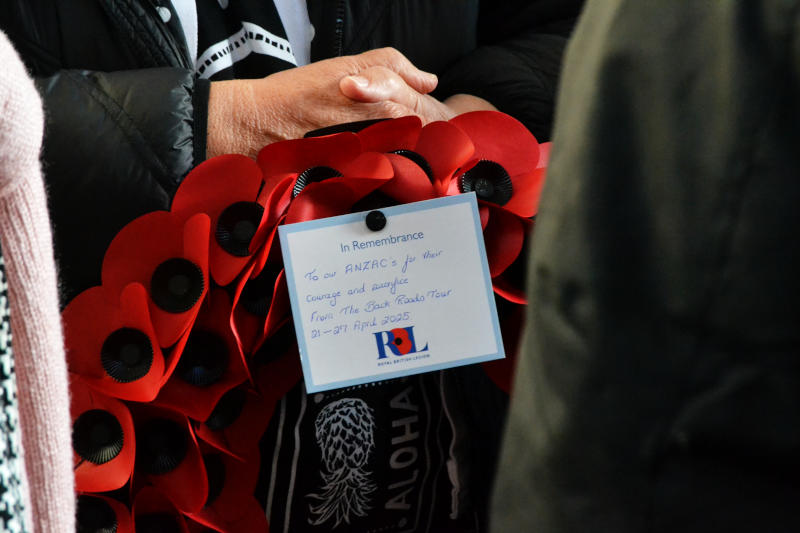 Folded hands above part of a wreath, on which a card is incribed: "In Remembrance: To our ANZAC's for their courage and sacrifice. From the Back Roads Tour 21-27 April 2025"
