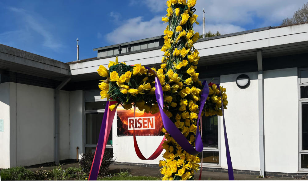 A cross covered with daffodils and a white building in the background
