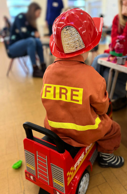 View from behind of a fire uniform and helmet: its wearer is sitting on a play fire engine