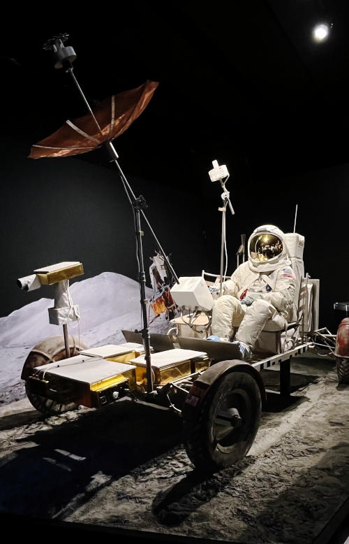 Model of a lunar rover from the Apollo programme with an astronaut sitting in one seat. The model is on a simulation of the lunar surface