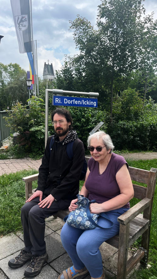 Martin and Miriam sitting on a bench by a roadside. Behind is a sign "Ri. Dorfen/Icking"