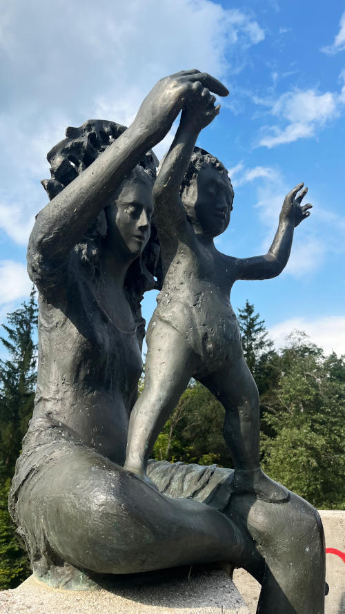 Madonna and Child statue made out of dark grey metal: Mary is playing with the infant Jesus who stands in her lap