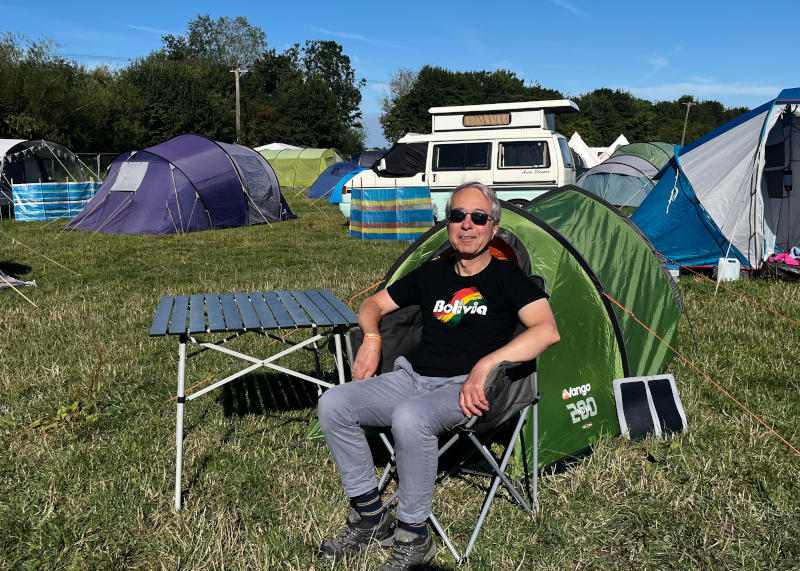 Phil sitting in a camping chair in front of a small green tent. A campervan and other, larger tents are in the background