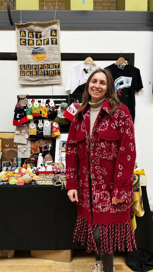 Iryna wearing a red coat standing in front of a stall selling a wide variety of items from Ukraine