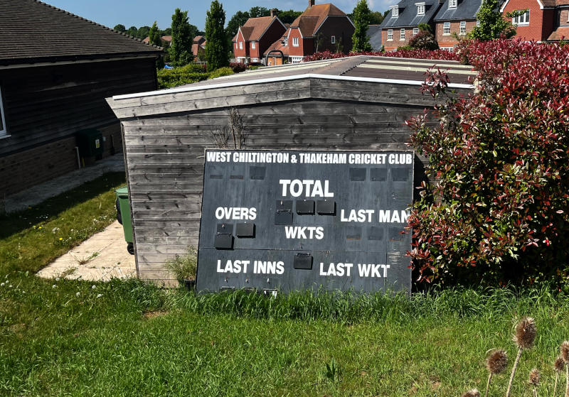 A small cricket scoreboard, the numbers all blank, lying on grass in front of a hut